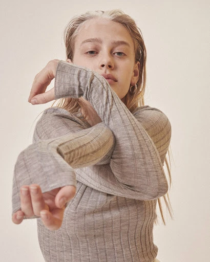 Blonde woman in ribbed grey top with long sleeves poses in a studio setting with neutral backdrop in a portrait orientation.