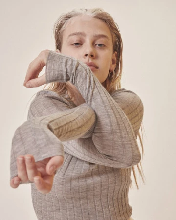 Blonde woman in ribbed grey top with long sleeves poses in a studio setting with neutral backdrop in a portrait orientation.