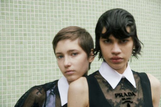 Two young fashion models in black lace tops and white collars pose against a square green tile background for a fashion editorial.