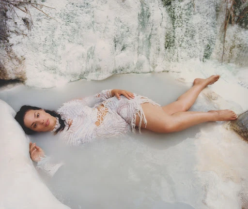 Woman in a white lace dress lies in pale blue thermal waters, surrounded by textured white rock formations, captured in a medium shot.