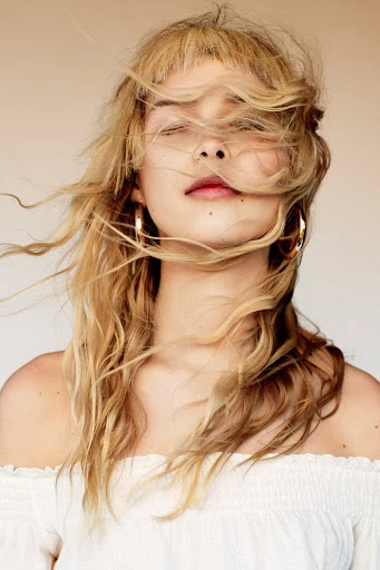 Close-up portrait of a blonde adult female with windblown hair covering her face, wearing gold hoop earrings and a white off-the-shoulder top.