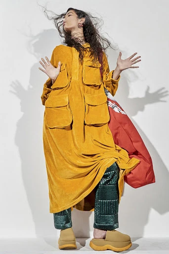 Full-length studio shot of a woman in an orange tunic and green pants, arms raised, accessorized with brown clogs and a red puffy jacket.