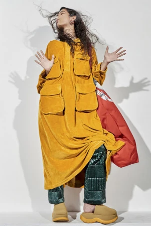 Full-length studio shot of a woman in an orange tunic and green pants, arms raised, accessorized with brown clogs and a red puffy jacket.