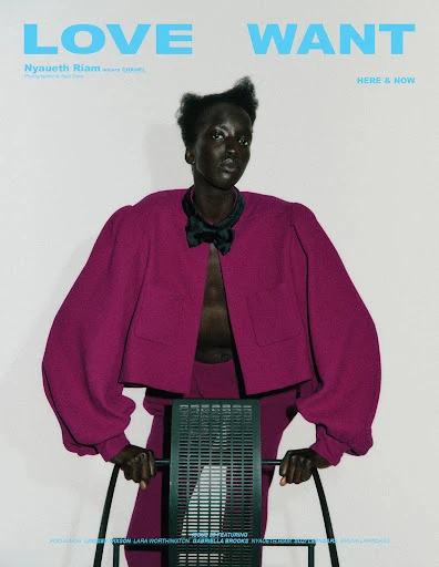 Fashion editorial shot of a Black model, Nyaueth Riam, wearing a magenta suit jacket and matching pants, posing with a metal chair.