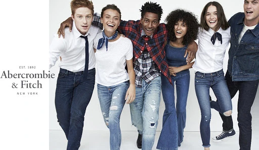 Group of six models with arms linked laughing together against a bright white background wearing denim and casual shirts.