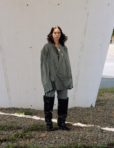Woman in an oversized grey jacket and black boots stands outside against a white wall and a gravel surface, under natural light.