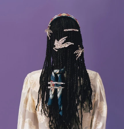 Woman with dark braided hair facing away, adorned with jeweled accessories and a patterned scarf, set against a soft purple backdrop.