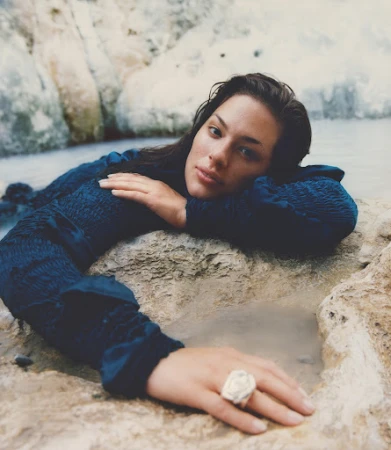 Woman in dark blue textured long-sleeve dress resting on light stone rocks by water, photographed by Marco Braca
