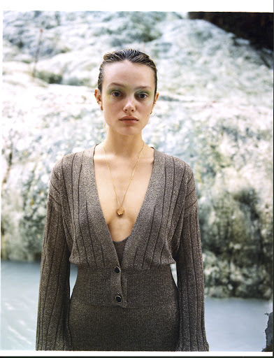 Portrait of a woman with green eye makeup, wearing a gold necklace and a brown, ribbed, V-neck dress, standing in front of white rocks and blue water.