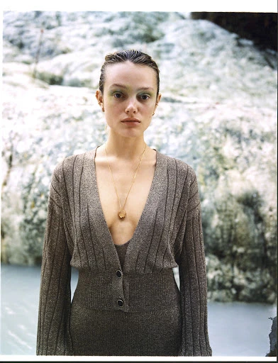 Portrait of a woman with green eye makeup, wearing a gold necklace and a brown, ribbed, V-neck dress, standing in front of white rocks and blue water.