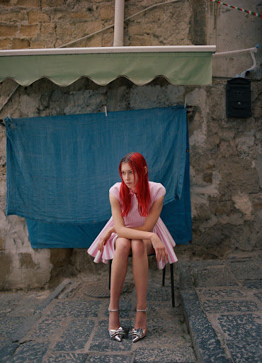 A young woman with vibrant red hair is sitting on a chair in an old town alley, posing for a fashion shoot in a pink dress and silver heels.