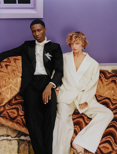 Stylish duo poses indoors: elegant Black man in tuxedo with white accents & curly blonde woman in white suit, sitting on patterned couch.