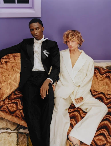 Stylish duo poses indoors: elegant Black man in tuxedo with white accents & curly blonde woman in white suit, sitting on patterned couch.