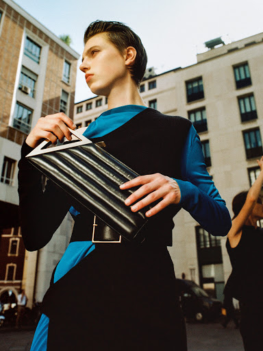 A woman in a black and blue dress holds a black clutch bag with a white triangular design in an urban setting with tall buildings.