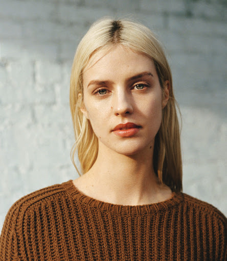 Close-up portrait of a blonde woman with blue eyes wearing a brown sweater against a white brick wall, showcasing a natural look.