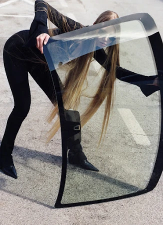 Young woman in black outfit poses with a clear car windshield on a gray asphalt surface, presenting a sleek and modern aesthetic.