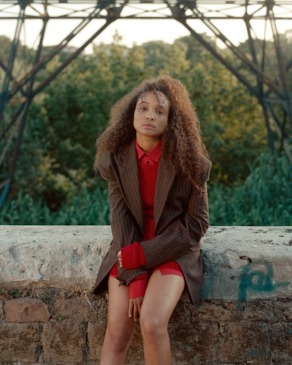 Portrait of a woman with curly hair in a blazer and red top, sitting on a ledge with a bridge and trees in the background.
