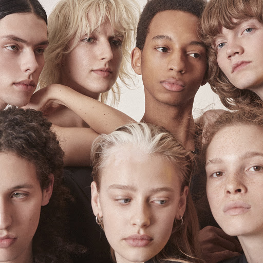 Diverse group of seven young adult models posing in a close-up studio shot, showcasing various skin tones and hair colors in a beauty or fashion context.