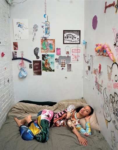 Young woman reclines on a bed in a room with walls adorned with artwork and personal items, creating a unique and intimate indoor setting.