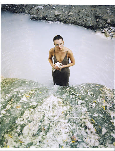 Woman in dress stands waist-deep in pale-blue water, holding a white bird, with rocky, earthy landscape elements visible.