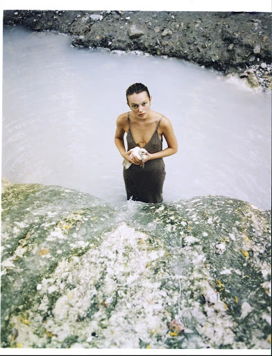 Woman in dress stands waist-deep in pale-blue water, holding a white bird, with rocky, earthy landscape elements visible.