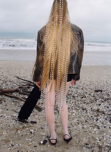 Woman with long blonde crimped hair and beaded extensions stands on a beach wearing a leather jacket, dark shorts, and light tights.