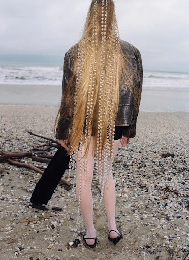 Woman with long blonde crimped hair and beaded extensions stands on a beach wearing a leather jacket, dark shorts, and light tights.