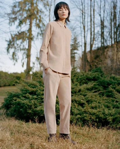 Adult woman stands in neutral-toned casual outfit in outdoor setting with grass, trees, and green bushes under a bright sky.
