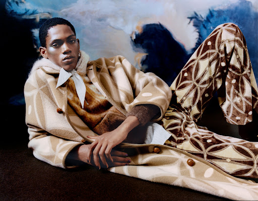 An adult Black male poses in patterned coat and pants against an abstract backdrop, evoking high fashion and creative studio portraiture.
