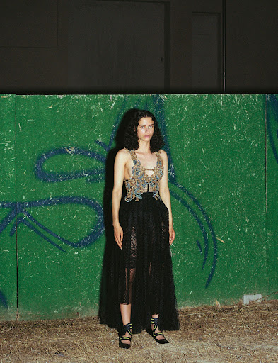 A stylish fashion photograph showcases a slender woman in a detailed top and black lace skirt against a textured green wall.