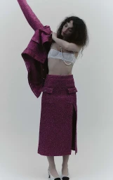 Woman with curly dark hair putting on a magenta tweed jacket over a white bra and matching skirt against a neutral background.