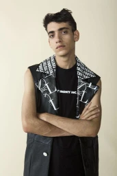 Young man with arms crossed wearing a black t-shirt and black leather vest against a plain neutral background.