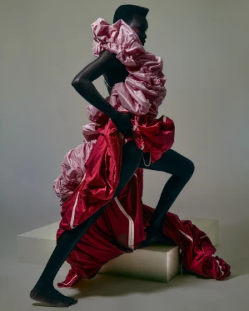 Model in voluminous pink ruffled top and red satin garment posing on a block against a grey studio backdrop