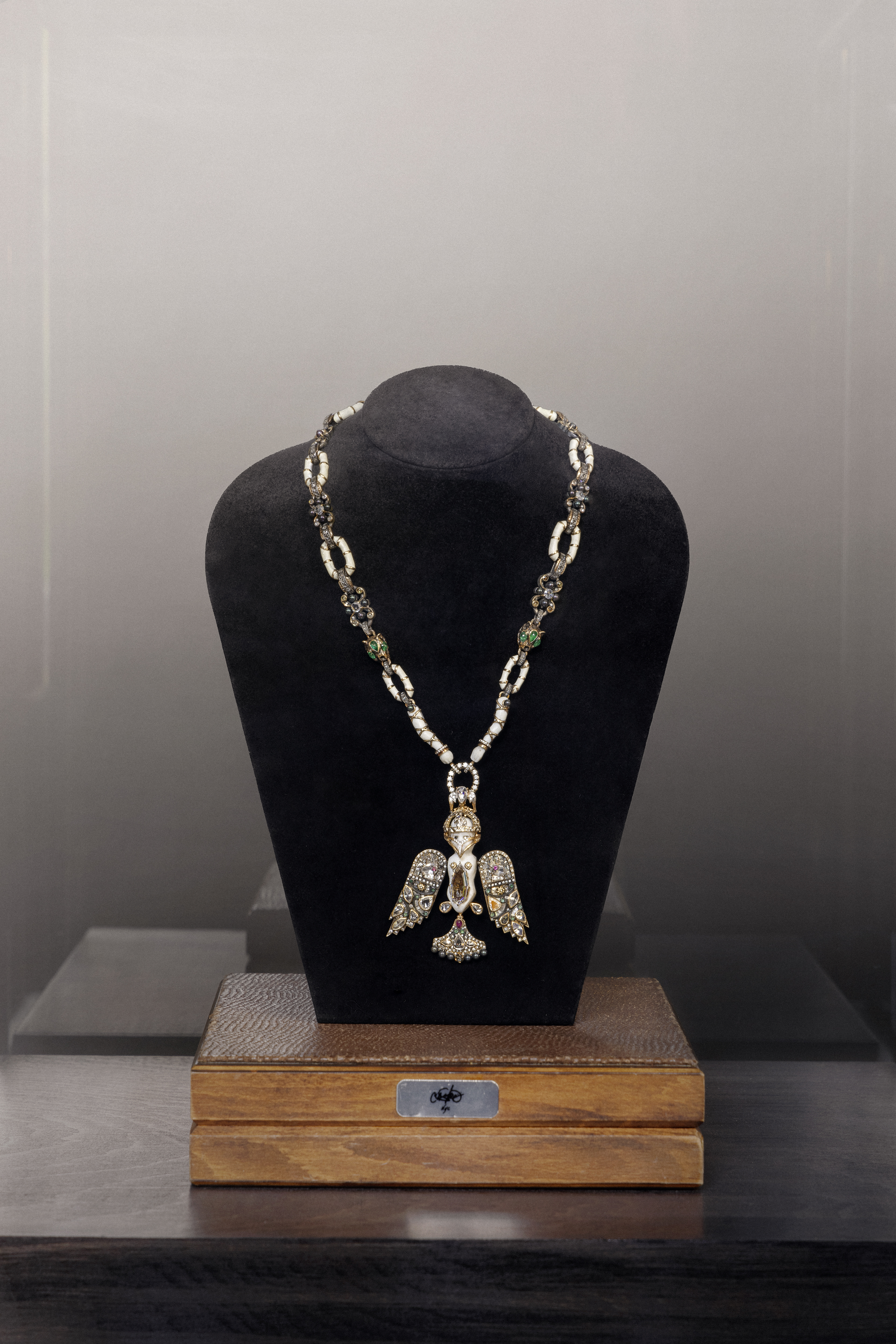 Elegant silver necklace with an avian-shaped pendant displayed on a black velvet bust on a wooden platform against a grey background.
