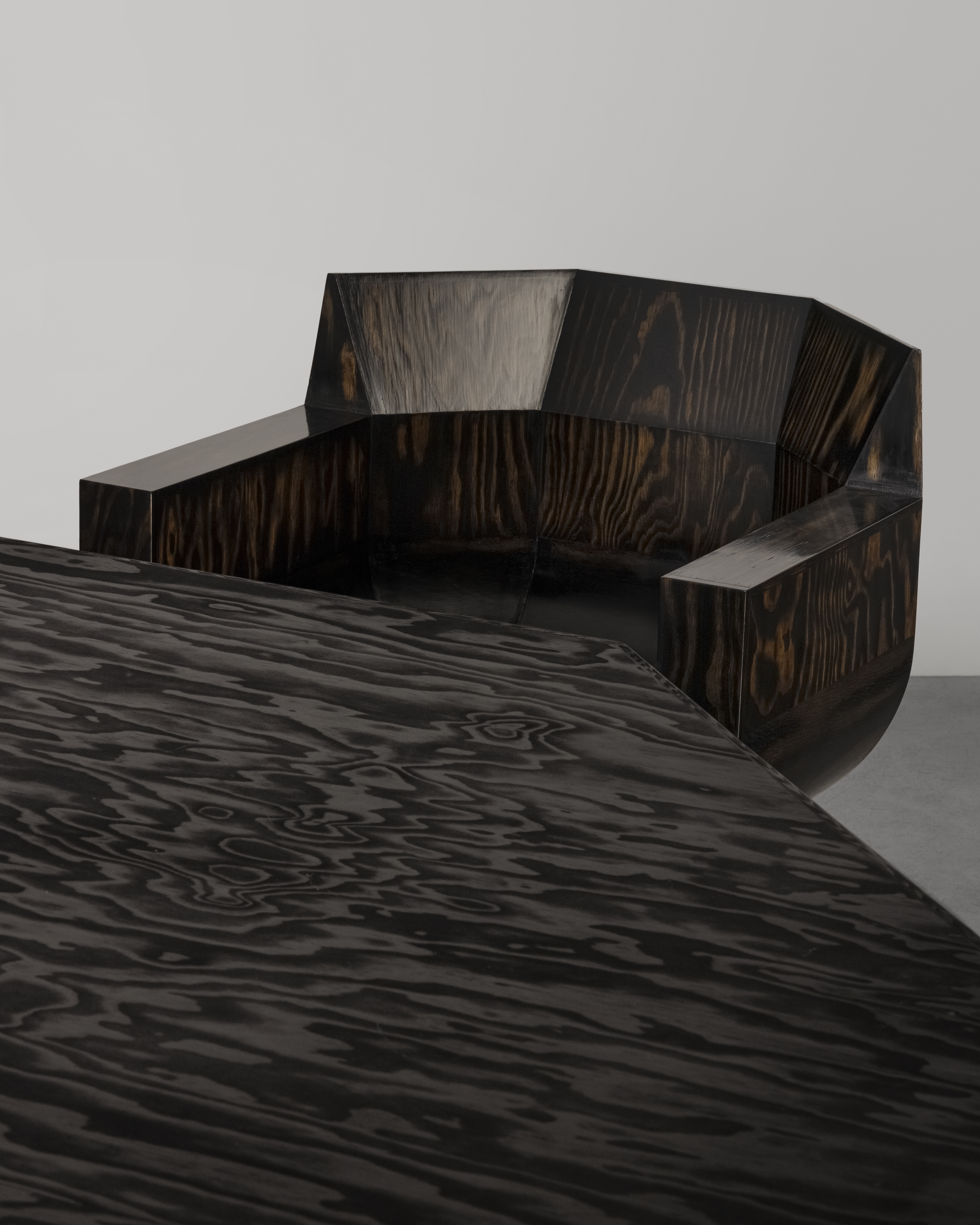 Geometric wood chair and table set with dark, swirling grain, highlighted by bright studio lighting against a light gray backdrop.