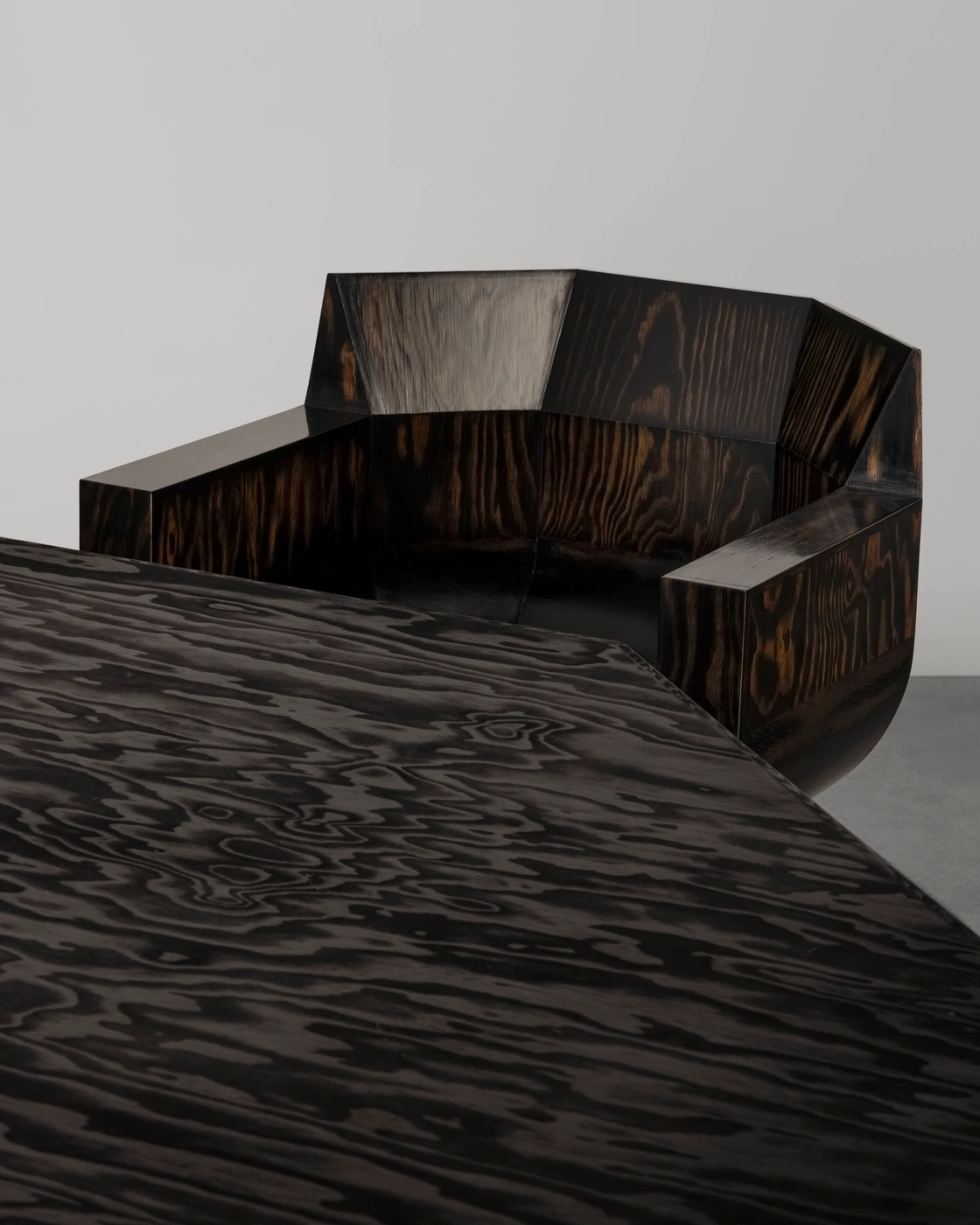 Sculptural dark stained wood armchair with a faceted backrest positioned behind a matching wooden table.