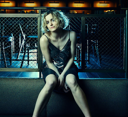 Blonde woman with metallic dress sits with crossed legs in a dimly lit indoor space, gazing into the camera with a serious expression.