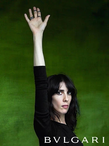Brunette woman poses with raised hand displaying rings against a green backdrop; Bulgari logo visible at the bottom of the frame.