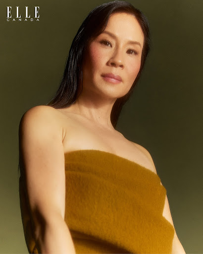 Close-up portrait of Lucy Liu, Asian woman with shoulder-length dark hair, wearing a mustard-yellow strapless top, against an olive-green background.