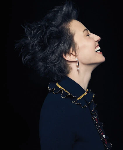 Elegant woman laughs in profile against a black background. She has short curly dark hair and wears a blue dress with gold and white trim.