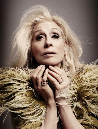 Portrait of an older woman with voluminous blonde hair wearing a feathered collar, hands raised to her chin with rings visible.