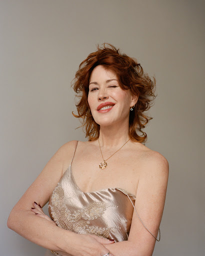 Red-haired woman with a slight smile and winking in front of gray background wearing a spaghetti strap dress and jewelry, portrait shot.