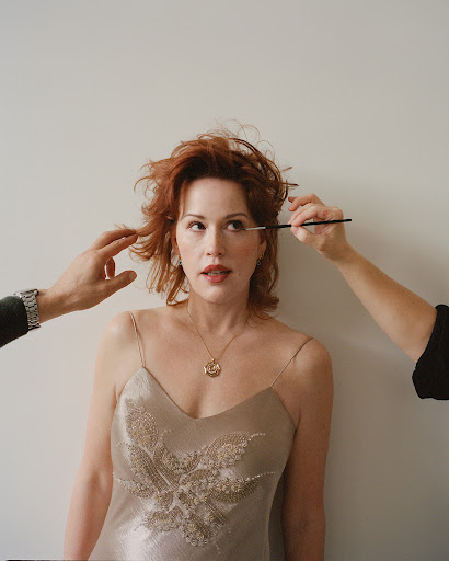 Adult woman with red hair getting makeup applied by two makeup artists. The woman wears a champagne colored dress with embroidery.
