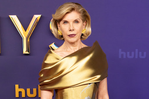 Close-up of Christine Baranski at an event, wearing a gold dress with dramatic shoulder and statement earrings, against a branded backdrop.