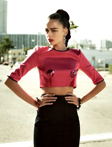 Fashionable adult female in pink crop top and black pencil skirt poses outdoors with an urban setting and buildings in the background.