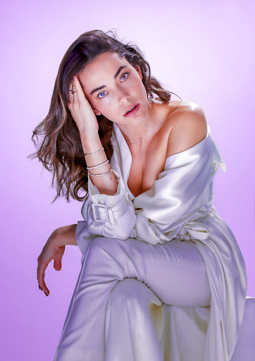 Elegant woman poses in a draped white outfit against a soft lavender background. She wears bracelets and a ring, and has long brunette hair.