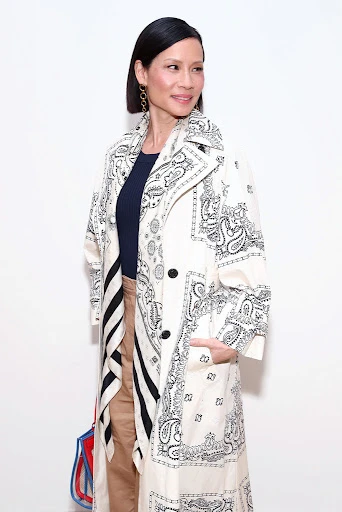 Adult female, Lucy Liu, wearing a paisley-patterned trench coat and holding a red, white, and blue tote bag in a photo studio setting.