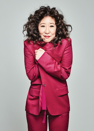 Attractive Asian woman with curly hair poses in a pink suit against a gray background. The image is a portrait-style medium shot.