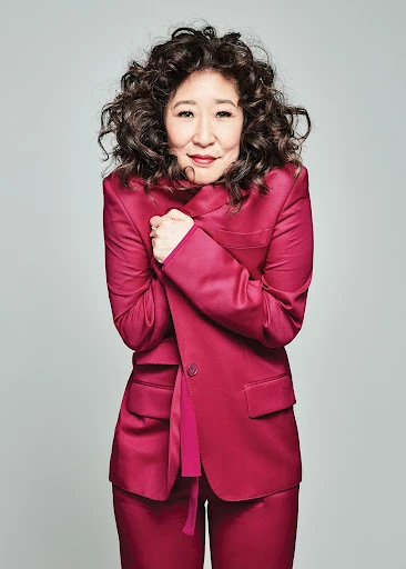 Attractive Asian woman with curly hair poses in a pink suit against a gray background. The image is a portrait-style medium shot.