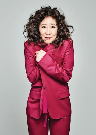 Attractive Asian woman with curly hair poses in a pink suit against a gray background. The image is a portrait-style medium shot.